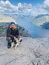 Person med liten hund sitter på fjellhylle med utsikt over fjord og fjell