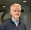 En mann i mørk genser står innendørs foran en vegg med delvis synlig logo.