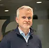 En mann i mørk genser står innendørs foran en vegg med delvis synlig logo.