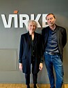 To personer står foran en grå vegg med stor logo.