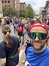Mange løpere står samlet i en bygate før start, med startnummer og en mann i forgrunnen.