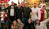 Smilende gruppe supportere står samlet foran tribunene på en fotballarena.
