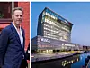 Et moderne glasskontorbygg ved vannet i skumringen, med en mann avbildet til venstre foran en rød murvegg.