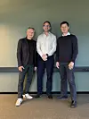 Tre menn står foran en nøytral vegg i et innendørs rom.