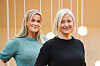 To kvinner står foran en lys vegg med hengende lamper i et varmt opplyst rom.