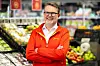 En butikkansatt i rød uniform står i en fruktavdeling med varer i bakgrunnen.