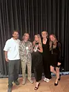 Fem personer poserer foran et svart sceneteppet i et innendørs rom med tregulv.