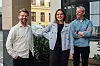 Tre personer står utendørs foran en bygning og ser mot kameraet.