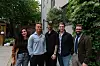 Fem personer står sammen utendørs i en gårdsplass med grønne planter og bygninger rundt.