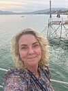 Kvinne tar selfie ved en brygge med stille vann og fjell i bakgrunnen.