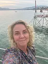 Kvinne tar selfie ved en brygge med stille vann og fjell i bakgrunnen.