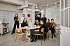 En gruppe mennesker samlet rundt et bord i et lyst studio med store vinduer og industrielle detaljer.