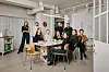 En gruppe mennesker samlet rundt et bord i et lyst studio med store vinduer og industrielle detaljer.