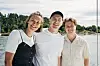 Tre personer står tett sammen og smiler ved vannkanten med trær og himmel bak.