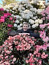 Smilende person stikker hodet opp blant hvite og rosa rhododendronblomster i en hage.