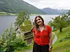 Kvinne i rød bluse står ved en sti med fjord, fjell og grønn natur i bakgrunnen.