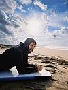 Person i svart våtdrakt ligger på et surfbrett på stranden under en lys himmel.
