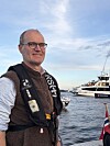 En mann står ved en havn med ferge og båter bak seg under en blå himmel.