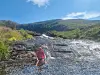 To personer står i grunt vann ved en steinete elv med fjell og blå himmel i bakgrunnen.