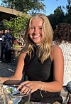 Smilende ung kvinne sitter ved et utendørs bord med glass og mobiltelefon.