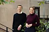 To personer står foran en moderne bygning med lyse vegger, vinduer og grønne planter.