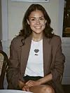 Smilende kvinne i brun blazer og hvit Alba-skjorte sitter på en stol innendørs.
