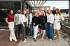 Gruppe voksne poserer på en takterrasse med glasstak, steinbed og utsikt mot vannet.