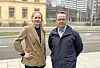 To personer står på et fortau foran bygninger og en vei i et urbant miljø.