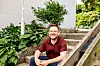 Smilende mann i rød skjorte sitter ved en trapp med grønne planter rundt seg.