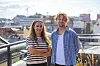 To smilende personer står på en takterrasse med byutsikt.