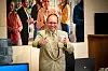 Mann i beige dress med kaffekopp foran kolleger på et kontor.