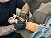 Tatoverer med hansker arbeider på kundens legg i en polstret stol.