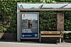Bussholdeplass med benk, glasskur og plakat foran en grønn hekk.