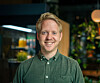 Smilende mann i grønn skjorte fotografert innendørs foran planter og varmt lys.