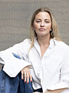 Kvinne med lyst hår i hvit skjorte og jeans, fotografert mot en gråmønstret bakgrunn.