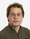 Mann med runde briller og olivengrønn genser fotografert mot lys bakgrunn.