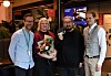 Fire personer poserer sammen innendørs, mens en av dem holder en blomsterbukett.