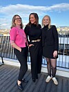 Tre kvinner står sammen på en terrasse med byutsikt og blå himmel.